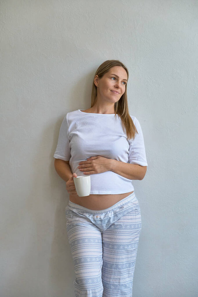 Pijamalı güzel hamile kadın evdeki beyaz bir duvarın arkasında duruyor. - Fotoğraf, Görsel