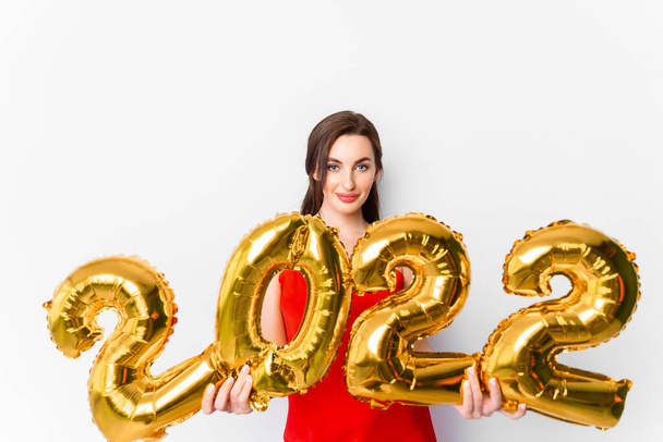 Young smiling woman in red cocktail dress with bright make-up celebrating New Year 2022 and holding golden balloons 2022 in hands on white background - Photo, Image