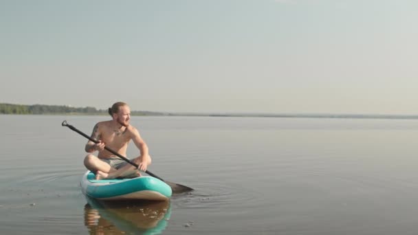 Hitaasti laaja kuva nuori mies istuu sup board melonta järvessä yksin aurinkoisena päivänä - Materiaali, video