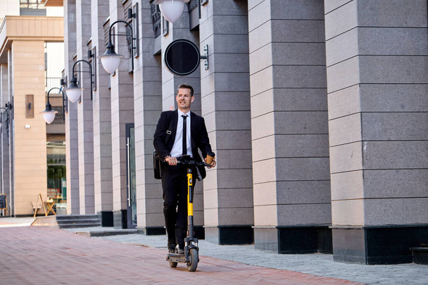 Optimistic caucasian business man in suit riding an electric scooter after business meeting - Fotó, kép