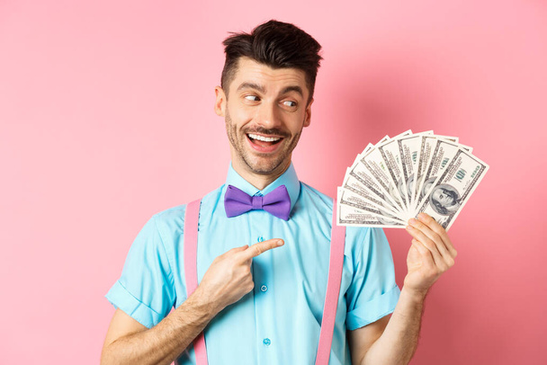 Reicher gutaussehender Mann in festlichem Outfit zeigt auf Geld, hält Dollars in der Hand und sieht glücklich aus, steht auf rosa Hintergrund - Foto, Bild