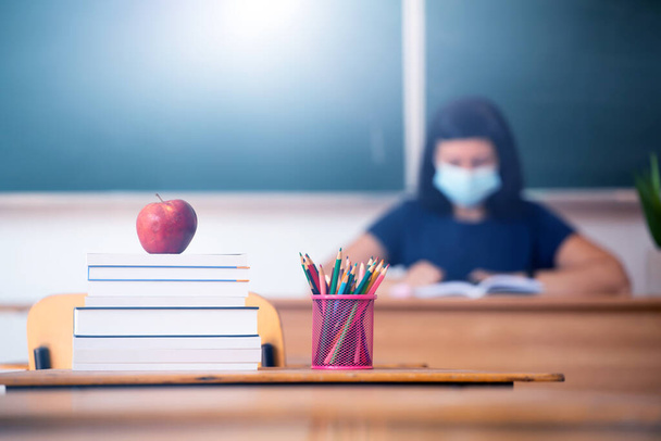 Back to School Concept. Apple, accessories and books in the classroom, teacher wearing mask in the background. - Photo, image