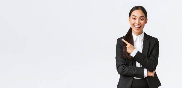 Excited attractive asian female saleswoman, real estate agent in suit suggesting perfect house, standing in suit and pointing finger left. Businesswoman making announcement, show banner - Foto, Imagen