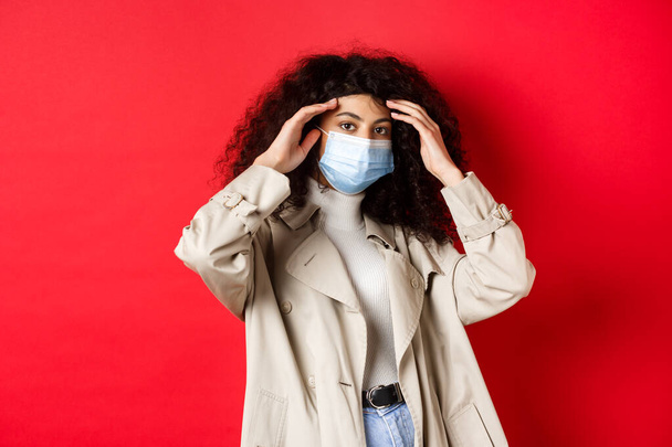 Covid-19, pandemická a karanténní koncepce. Stylová mladá žena s kudrnatými vlasy, jít ven v lékařské masce a trenč kabát, opravit sestřih, stojící na červeném pozadí - Fotografie, Obrázek