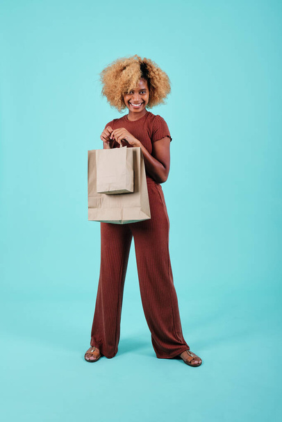 Afro vrouw glimlachend terwijl het houden van boodschappentassen op een lichtblauwe geïsoleerde achtergrond. Black Friday verkoopseizoen concept. - Foto, afbeelding
