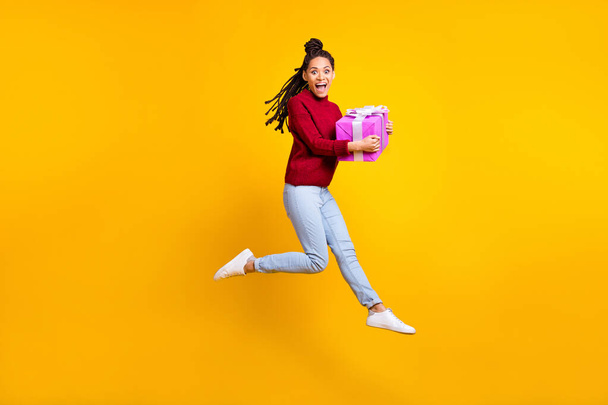 Full length photo of charming cute dark skin lady wear red pullover smiling jumping giving present isolated yellow color background - Photo, image