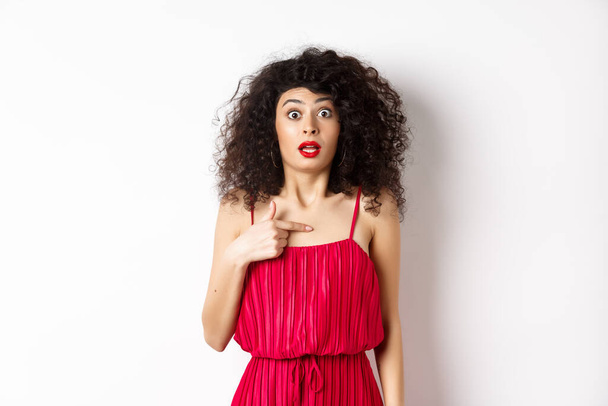 Überraschte junge Frau in rotem Kleid, die verwirrt und schockiert auf sich selbst zeigt, steht auf weißem Hintergrund - Foto, Bild