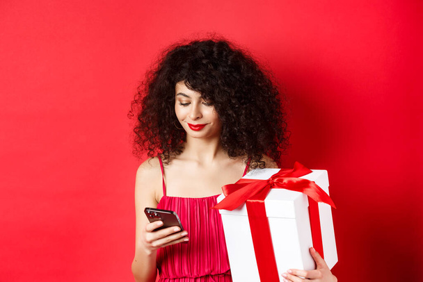 Ημέρα του Αγίου Βαλεντίνου και ψώνια. Όμορφη καυκάσια γυναίκα με κόκκινο φόρεμα, κρατώντας κουτί δώρου από τον εραστή και χρησιμοποιώντας το κινητό τηλέφωνο, ανάγνωση μήνυμα στο smartphone, φόντο στούντιο - Φωτογραφία, εικόνα