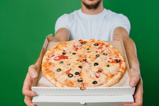 Nuori mies valkoisessa t-paidassa pitää pizzalaatikkoa käsissään vihreällä taustalla. Komea kaveri Pizza toimitus kaveri - Valokuva, kuva
