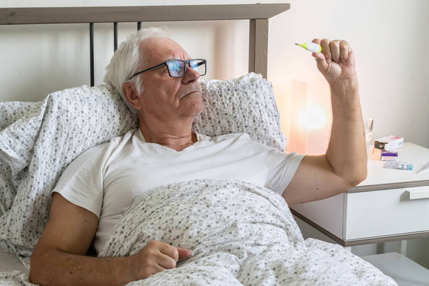 aging senior man male in bed at home flat tired sick ill alone retired resting virus taking care unhappy sad, holding thermometer - Foto, afbeelding