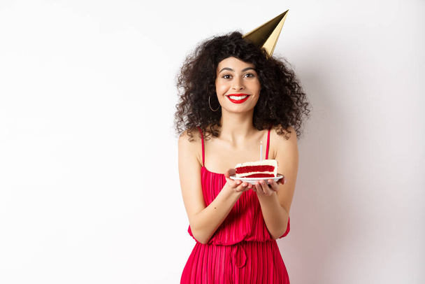 Beautiful woman in red dress, wearing party hat and celebrating birthday, holding b-day cake and making wish, smiling at camera, standing on white background - Photo, Image