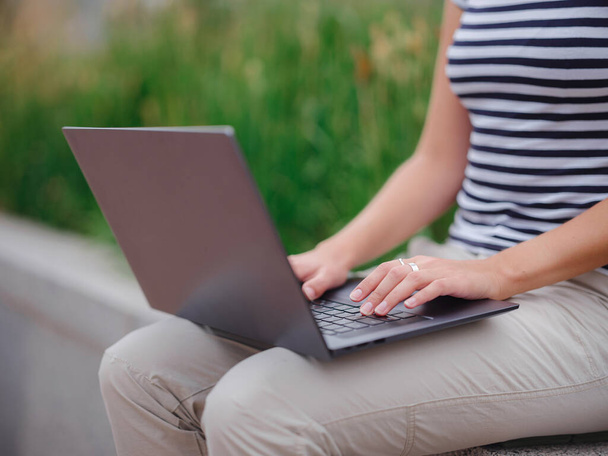 szczęśliwa azjatycka studentka pracująca z laptopem w nowoczesnym kampusie uniwersyteckim. Kobieta w koszulce w paski i lekkie spodnie pracuje w szarym laptopie. Ręce do góry. - Zdjęcie, obraz