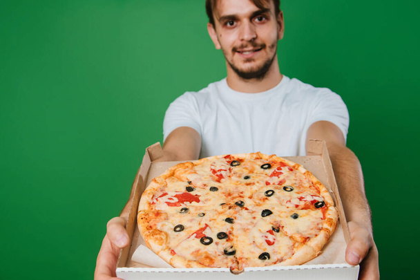 Un joven con una camiseta blanca sostiene una caja de pizza en sus manos sobre un fondo verde. Chico guapo Repartidor de pizza - Foto, imagen