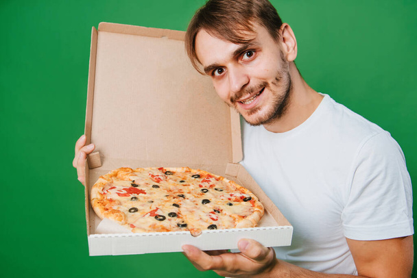 A young man in a white T-shirt holds a pizza box in his hands on a green background. Handsome guy Pizza delivery guy - Zdjęcie, obraz