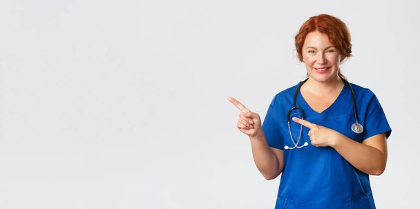 Gyógyszer, egészségügy és koronavírus koncepció. Kellemes női orvos portréja, piros hajú ápolónő kék köpenyben, aki vidáman néz ki, mutatja a klinika plakátját, kórházi hirdetést, mutogató ujjal balra - Fotó, kép