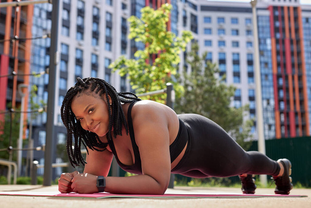 Smiling african overweight female doing plank on fitness mat outdoors, side view portrait - Φωτογραφία, εικόνα