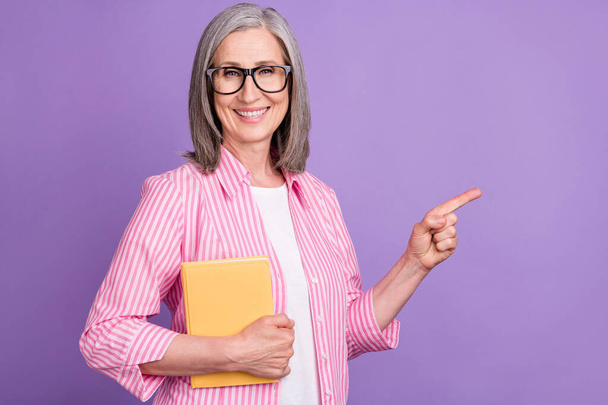 Photo of mature woman happy smile hold book point finger empty space advertise recommend isolated over purple color background - Zdjęcie, obraz