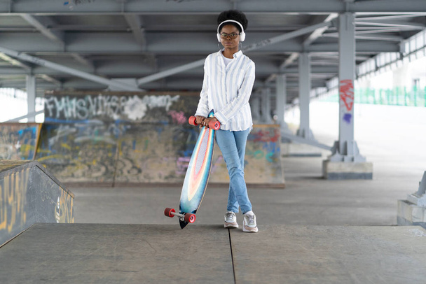 African girl with skateboard. Trendy female dressed in urban outfit wear headphones, hold longboard - Photo, image
