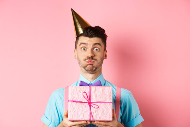 Vacaciones y concepto de celebración. Chico divertido mirando a la cámara sorprendida, usando sombrero de fiesta, sosteniendo regalo de cumpleaños y conteniendo la respiración, haciendo pucheros en la cámara, fondo rosa - Foto, imagen