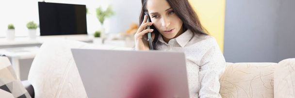 Молода жінка розмовляє по телефону і працює на ноутбуці, сидячи на дивані
 - Фото, зображення