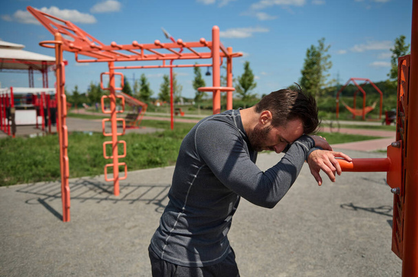 Вымотавшийся евроатлет, прислонившись лбом к перекладине, отдыхает после тяжелой тренировки на открытом воздухе на летней спортивной площадке - Фото, изображение