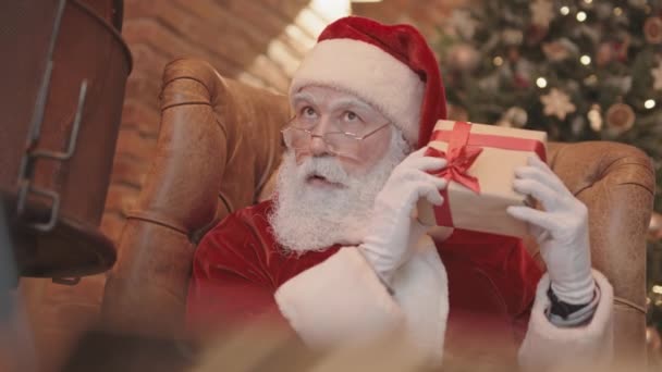 Niski kąt świętego Mikołaja siedzącego w fotelu w domu, trzęsącego się małym pudełkiem z prezentami i słuchającego, jak wydaje dźwięk, a następnie wyrzucającego go i patrzącego w kamerę - Materiał filmowy, wideo