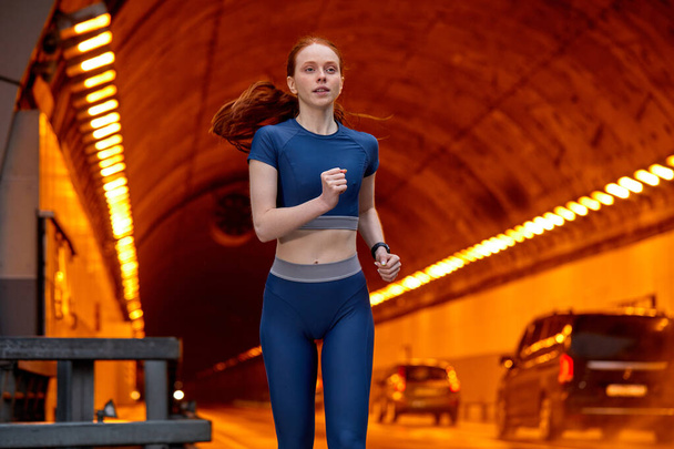 Athlete woman runner in blue top and leggins confidently running along the road - Zdjęcie, obraz