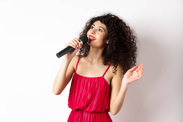 Όμορφη κυρία με κόκκινο φόρεμα τραγουδώντας τραγούδια στο μικρόφωνο, χαμογελώντας και κοιτάζοντας προς τα πάνω, στέκεται σε λευκό φόντο - Φωτογραφία, εικόνα