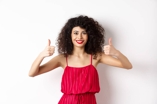 Pretty female model with curly hair and red dress, bright makeup, showing thumbs up in approval, say yes, smiling and nodding in approval, white background - Photo, Image