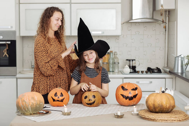 Gelukkige familie: moeder en dochter vieren Halloween. Vrolijk kind in heksencarnaval kostuum in de kamer. Vrolijk kind en ouder spelen thuis met pompoenen en zwarte heksenhoeden - Foto, afbeelding