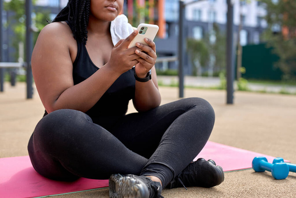 Vágott fénykép túlsúlyos afrikai nő ül Fitness matrac segítségével okostelefon - Fotó, kép