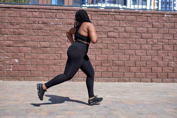 Overweight young african woman jogging outdoors. Fat beautiful female in black tracksuit - Zdjęcie, obraz