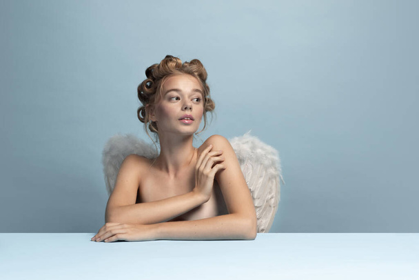 Portrait of young beautiful girl in image of angel with wings sitting and ogles isolated on blue gray studio background. Concept of beauty, purity, tenderness - Photo, Image