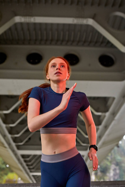Beautiful confident sports woman in stylish sportswear jogging on bridge - Photo, Image