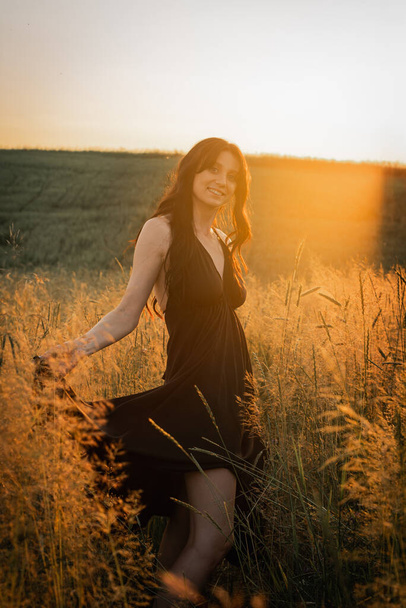 Sunset, soft golden evening colors, young, beautiful, emotional brunette girl with long hair in a black long dress on a golden wheat field. Cosiness of soul, joy, nature - Photo, Image