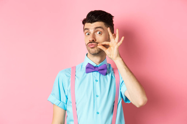 Lustiger junger Mann mit französischem Schnurrbart und Puckerlippen, der albern in die Kamera schaut, mit Fliege und Hosenträgern auf rosa Hintergrund - Foto, Bild