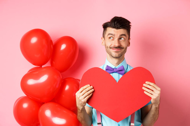 Valentines day concept. Smiling handsome man looking dreamy left, waiting for true love girlfriend with big red heart cutout, standing over pink background - Photo, Image