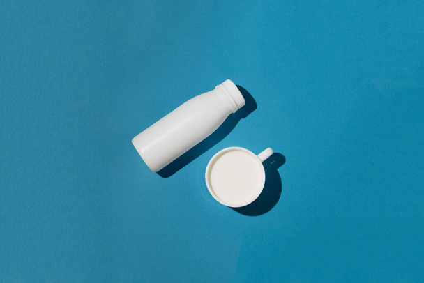 Bílá láhev a bílý šálek s mlékem nebo mléčným výrobkem na modrém pozadí, dieta a výživa koncept, horní pohled - Fotografie, Obrázek