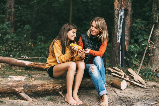 Una adolescente con su madre está sentada en un tronco en el bosque, una chica está comiendo maíz, haciendo senderismo el fin de semana - Foto, Imagen