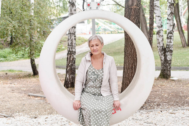 Portret van een droevige blanke senior vrouw in jurk met kort grijs haar zittend op een ronde bank in de zomer stadspark. - Foto, afbeelding