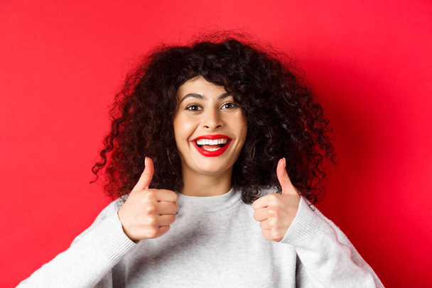 close-up van vrolijke blanke vrouw tonen duimen omhoog in goedkeuring, glimlachen blij, als en lof iets goeds, staande op rode achtergrond - Foto, afbeelding