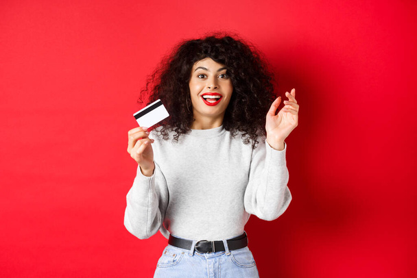 Nadšený žena vypráví o speciální nabídku, ukazuje plastovou kartu a vypadá šťastně, jít nakupovat, stojí na červeném pozadí - Fotografie, Obrázek