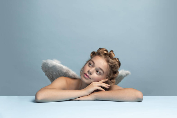 Dreaming angel, young beautiful girl sitting and thinking about cute moments of life isolated on blue gray studio background. Concept of beauty, purity, tenderness - Valokuva, kuva