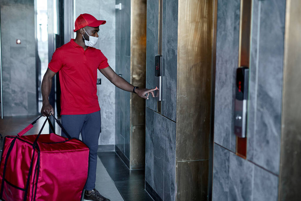 Afro american Male courier with thermo bag pushing elevator button,side view. - Фото, изображение