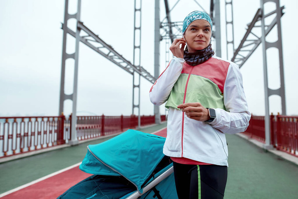 Sportive woman wearing earbuds while standing on the bridge with a baby carriage, ready for morning run on a cloudy day in the city - Foto, Imagem