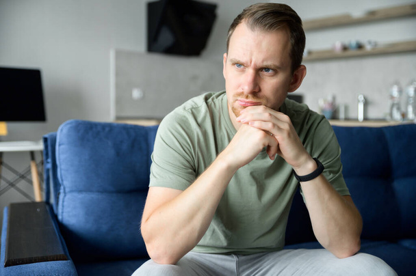 Worried guy sits on the sofa alone and looks away lost in thoughts - 写真・画像