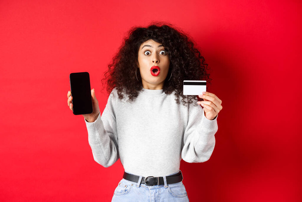 Excited woman with credit card showing empty smartphone screen, gasping amazed, standing against red background - Photo, Image