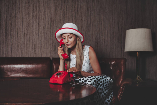 Portrait emotion adult woman actress with old red telephone posing dressed retro styling clothes in vintage living room. Female in pin-up fashion style black and white dress in polka dot and hat - Foto, Bild