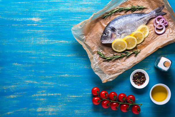 Raaka Dorado kala valmistetaan ruoanlaittoon viipaleiksi sitruunaa, rosmariinia, sipulia, kirsikkatomaattia, mausteita ja öljyä sinisellä pohjalla. Huippunäkymä. Kopioi tila. - Valokuva, kuva