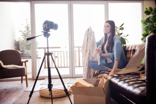 Portrait of attractive cheerful girl sitting on divan recording vlog video choosing buying clothes unpack order at home house indoors - 写真・画像